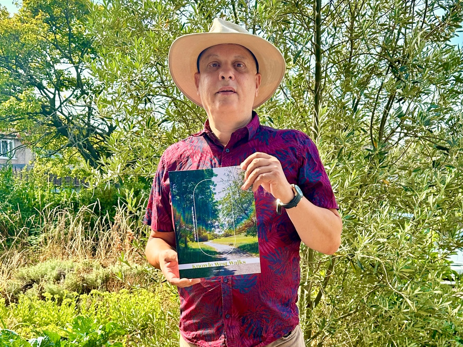 image of a man wearing a hat and holding and A4 sized book. He is standing in front of a tree